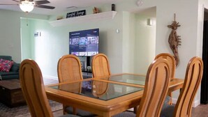 A dining area with a glass-top table and wooden chairs, a ceiling fan, a TV mounted on the wall, and a decorative wall piece.