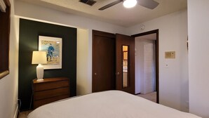 A bedroom with a bed, a nightstand with a lamp, a framed picture on the wall, and a door leading to another room.