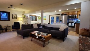 A modern living room with a dark-colored sofa, a coffee table, and a dining area with a small table and chairs. A kitchen with stainless steel appliances is visible in the background.