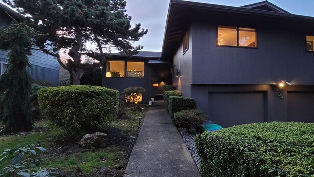 A modern two-story house with a dark exterior, a visible garage, and a well-maintained garden with hedges and a tree.