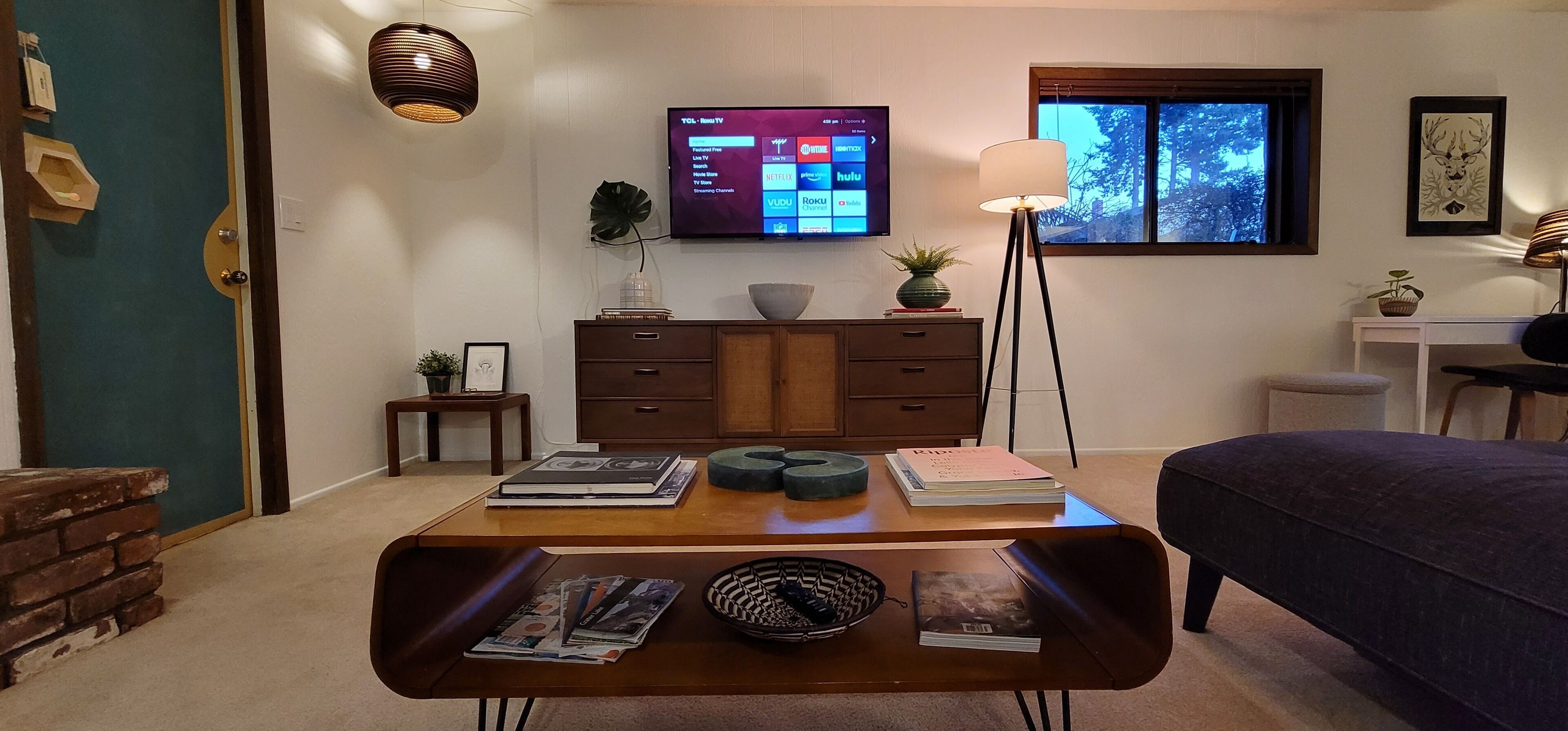 Une pièce avec un téléviseur fixé au mur, un bureau avec des livres et une lampe, une fenêtre donnant sur un beau paysage et un lit.