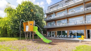 Un edificio moderno con varios balcones, un parque infantil con tobogán y un gran árbol al fondo.