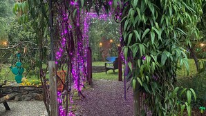 Um jardim com pérgola de madeira coberta por folhagem verde, luzes pendentes roxas e um caminho de pedra.