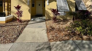 A two-story yellow building with white shutters, a covered entrance, and a small garden area with mulch and plants. - 5 minues to the theme park. Family sized communy pool (Kissimmee)