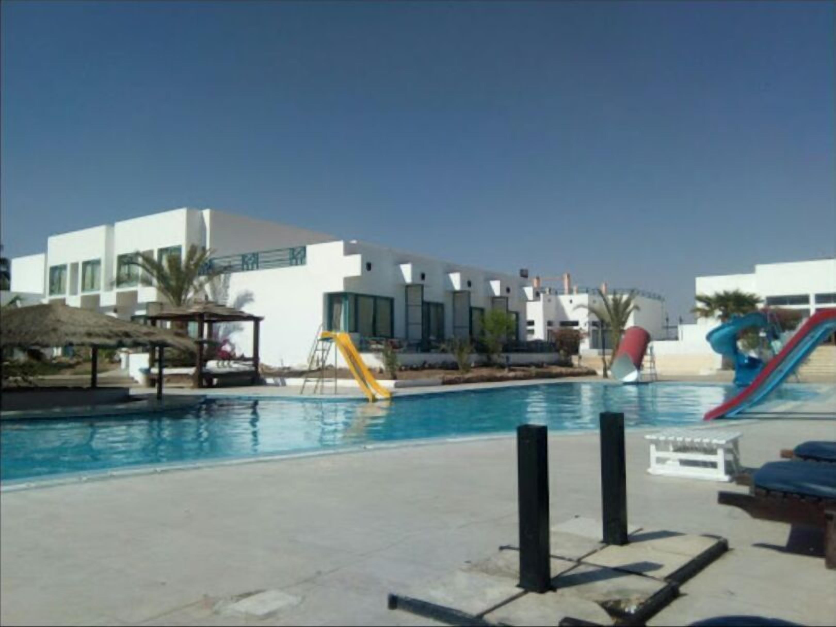 A swimming pool with slides, a thatched-roof structure, and modern buildings in the background.