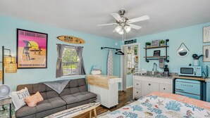 A room with a grey sofa, a bed with a beach-themed bedspread, a ceiling fan, a kitchenette with a blue appliance, and a surfboard sign on the wall. - Tiny Home Near the Bay by Beach's, Pensacola, Foley, Gulf Shores, Orange Beach
 (Elberta)
