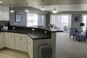 A modern kitchen with white cabinets, a black countertop, and a ceiling fan. There is a dining area with a patterned chair and a small table.