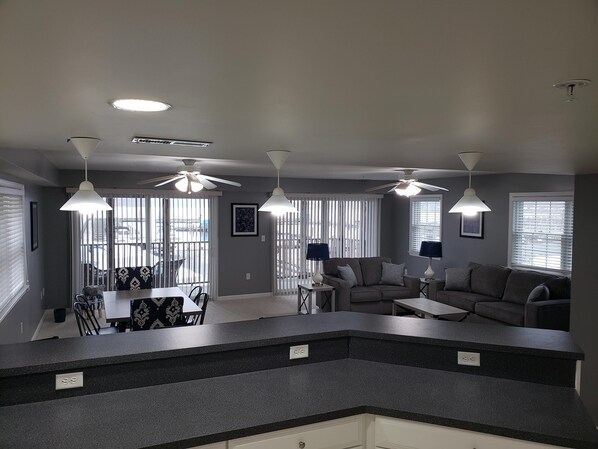 A modern kitchen with a dark countertop, a dining area with a table and chairs, and a living area with a sofa and two lamps.