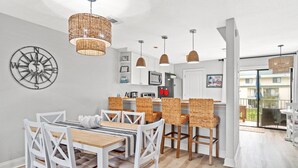 A modern kitchen with white cabinetry, a dining area with white chairs, and a large window overlooking a balcony. - Steps to beach Pool Parking 2 king beds beach gear Patio WI-FI (Miramar Beach)