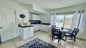 A compact kitchen with white cabinets, a microwave, and a stove. There's a dining area with a table and chairs, and a window with curtains. - Beach Tide Motel - #5 Studio 1 King bed (Hollywood)