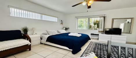 A bedroom with a bed, a sofa, a TV, a ceiling fan, and a window.