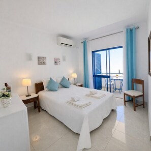 A hotel room with a large bed, two bedside tables with lamps, a chair, a balcony with a table and chair, and a view of the sea.