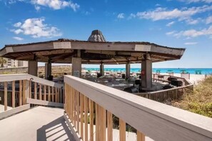 Ein hölzernes Gazebo mit Kegeldach, das einen Strandbereich mit Liegestühlen und Tischen überblickt.