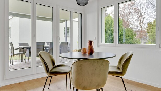 Ein Zimmer mit einem runden Tisch, vier Stühlen, großen Fenstern und Blick ins Freie.