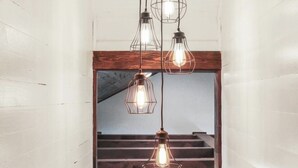 A narrow hallway with white wooden walls and a wooden ceiling. Three hanging pendant lights with exposed bulbs are visible. A doorway leads to a room with a wooden ceiling and a painting on the wall.