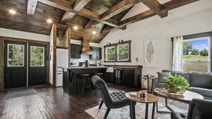 A spacious living area with a kitchen, wooden ceiling beams, and a large window offering a view of the outdoors.