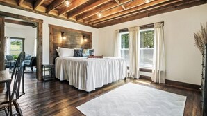 A bedroom with a wooden ceiling, a bed with white bedding, a large window with curtains, and a view of the outdoors.