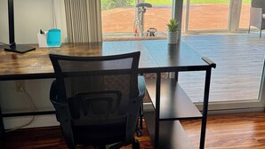 A modern office space with a wooden desk, a black office chair, and a view of a garden and lake through large windows. - Water front, river views, fishing from backyard, Large yard to enjoy. (Hampton)
