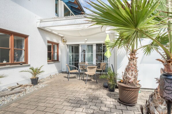 Une terrasse avec une table et des chaises, des plantes en pot et un bâtiment blanc doté de fenêtres et d’une porte.