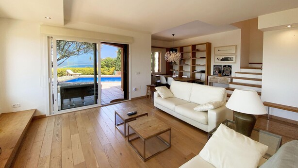 A spacious living room with a white sofa, a coffee table, and a bookshelf. A sliding glass door leads to a pool area with a view of the ocean. - Holiday rental villa with swimming pool in Begur, Centre (Begur)