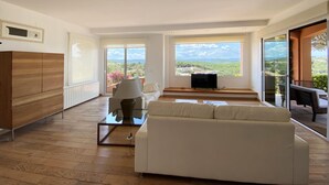 A spacious living room with a white sofa, a glass coffee table, and a TV on a stand. Large windows offer a view of a green landscape and mountains. - Holiday rental villa with swimming pool in Begur, Centre (Begur)