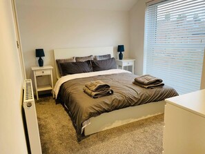 Un lit bien fait, avec une couette et des oreillers marron, une table de chevet blanche avec une lampe, et une fenêtre avec des stores.