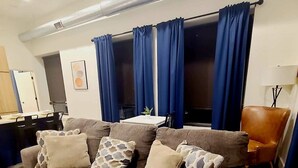 Ein Wohnzimmer mit braunem Sofa, gemusterten Kissen, einem blauen Vorhang, einem braunen Ledersessel und einem Teppich mit Muster.