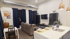 Ein modernes Wohnzimmer mit einer Couch, einem Esstisch mit Stühlen, einer Kücheninsel mit Spüle und einem flachbildfernseher an der Wand.