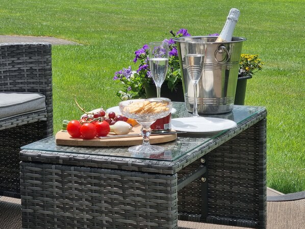 An outdoor dining setup with wicker furniture, a glass table, a bottle of champagne, a bowl of crackers, a plate of tomatoes, and a glass of water.
