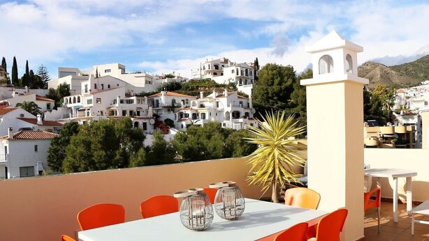 Eine Terrasse mit Tisch und Stühlen, einer Zierpflanze und Blick auf eine weiß getünchte Stadt mit Häusern und Bäumen.