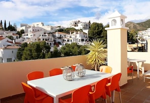A terrace with a table and chairs, a decorative plant, and a view of a white-washed town with buildings and trees.