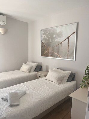Une chambre avec deux lits, une table de chevet, un climatiseur fixé au mur et un tableau représentant un pont de corde.