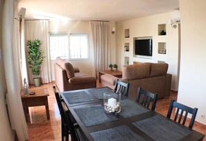 Un salon comprenant un canapé, une table à manger avec des chaises, un téléviseur fixé au mur et une fenêtre avec des rideaux.