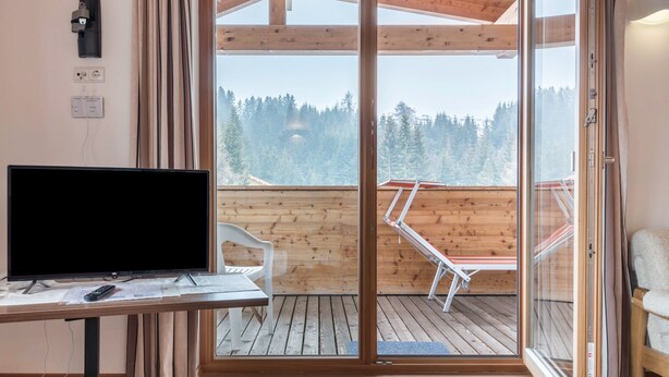 Ein Zimmer mit Fernseher auf einem Schreibtisch, einem Sessel, Holzboden und einer Glastür, die auf einen Balkon mit Blick auf Bäume führt.