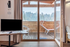 Una habitación con televisión en un escritorio, una silla, piso de madera y un ventanal de vidrio que da a un balcón con vistas a árboles.
