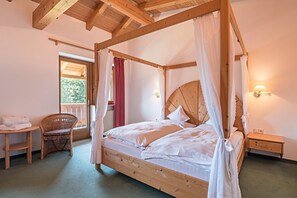 Ein Bett mit Baldachin, ausgestattet mit weißen Leinentüchern, ein Korbstuhl, ein Nachttisch mit Lampe sowie Blick ins Freie durch ein Fenster.
