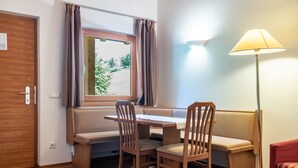 Um quarto com piso de madeira, janela com cortinas, mesa com cadeiras, sofá, luminária e porta.