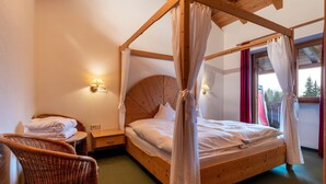 Ein hölzernes Himmelbett mit weißer Bettwäsche, ein Korbstuhl, ein Nachttisch und ein Fenster mit Vorhängen.
