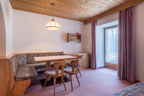 A room with a wooden ceiling, a dining table with chairs, a bench, a hanging light fixture, a window with curtains, and a small shelf with items.