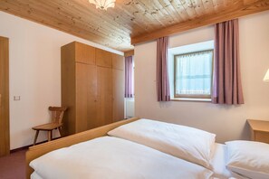 A room with a wooden ceiling, a window with curtains, a bed with white bedding, a wooden wardrobe, and a chair.