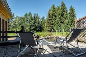 Two outdoor chairs facing a small pond with a wooden deck and a building in the background.
