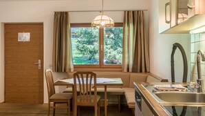 A kitchen with a dining area, a window with curtains, a wooden door, and a view of greenery outside.
