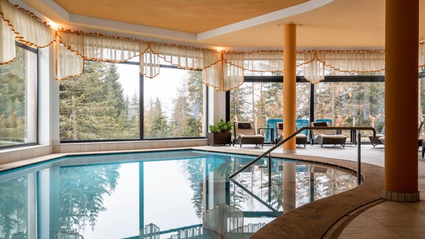 An indoor swimming pool with clear glass walls, large windows, and a view of trees outside.