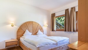 Una cama con estructura de madera y ropa de cama blanca, dos mesitas de noche con lámparas, una ventana con cortinas y vista a la vegetación exterior.