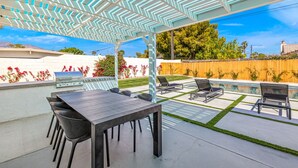 Un patio exterior con una parrilla, una mesa, sillas, una piscina y un área cubierta con techo geométrico.