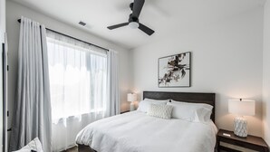 Une chambre à coucher bien aménagée, avec un grand lit, deux tables de chevet avec des lampes, un ventilateur de plafond et une fenêtre ornée de rideaux légers.
