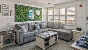 Un salon moderne avec un canapé d’angle gris, une table basse en bois et un mur vert orné d’une fresque sur le thème du surf.