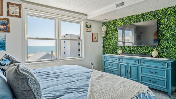 Une chambre avec une grande fenêtre, un lit, une commode et un mur vert orné d’un miroir.