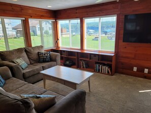 A cozy living room with a brown leather sofa, a white coffee table, and a wooden bookshelf filled with books. Large windows offer a view of a green lawn and a building in the distance. - Owasco Lake Finger Lakes Getaway (Moravia)