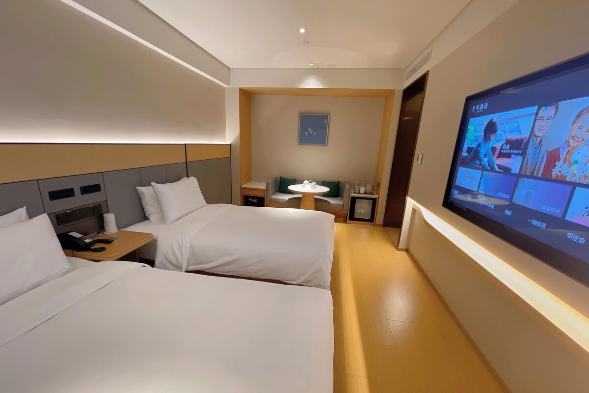 A hotel room with two beds, a TV, a small table with a chair, and a nightstand with a phone and cup.